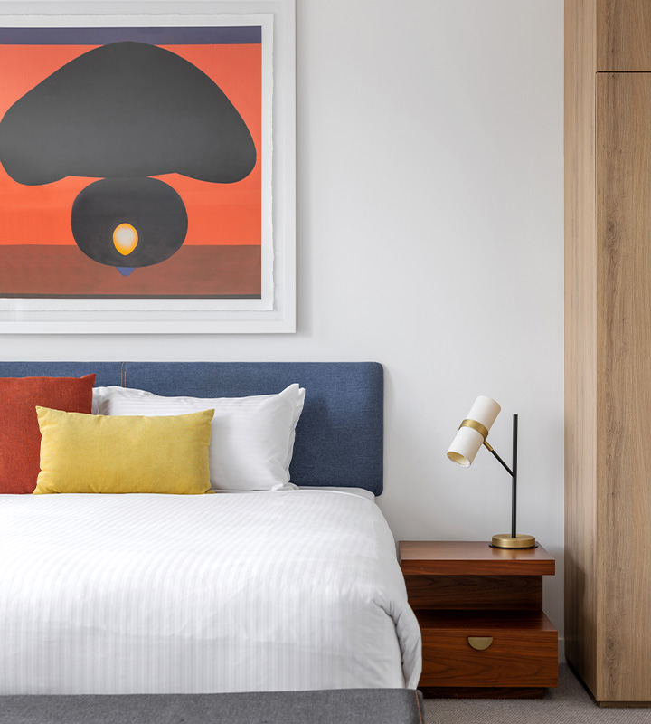 A hotel bed with yellow and red pillows and an artwork above the bed